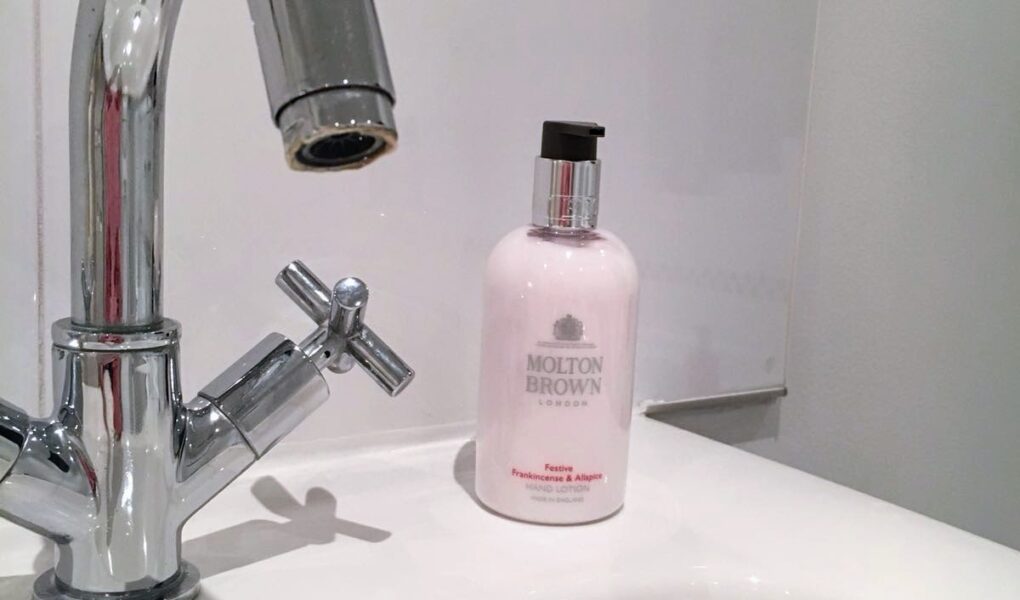 A chrome bathroom faucet is positioned over a white sink. Next to the faucet sits a pink bottle of Molton Brown hand lotion. The background features white tiles with a black and white checkered border near the top.