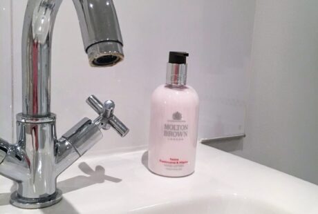 A chrome bathroom faucet is positioned over a white sink. Next to the faucet sits a pink bottle of Molton Brown hand lotion. The background features white tiles with a black and white checkered border near the top.