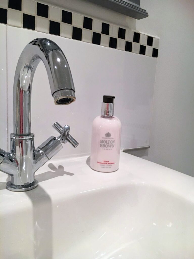 A chrome bathroom faucet is positioned over a white sink. Next to the faucet sits a pink bottle of Molton Brown hand lotion. The background features white tiles with a black and white checkered border near the top.
