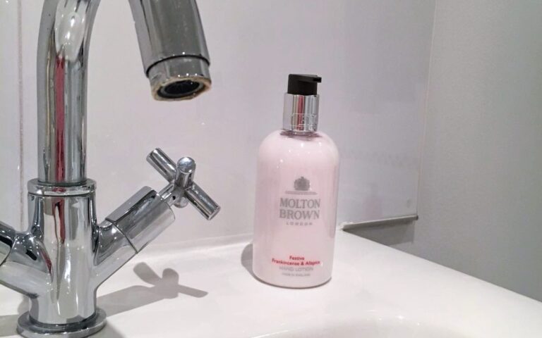 A chrome bathroom faucet is positioned over a white sink. Next to the faucet sits a pink bottle of Molton Brown hand lotion. The background features white tiles with a black and white checkered border near the top.