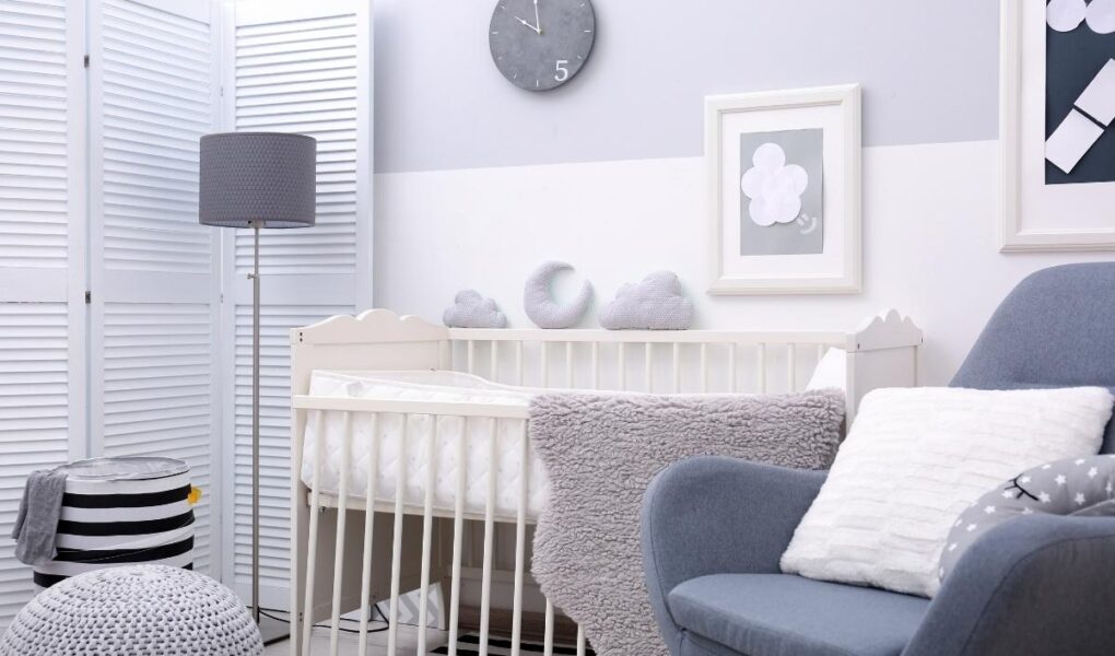A cozy, modern nursery with a white crib, grey cushions shaped like clouds, a grey armchair with a white pillow, a fluffy grey blanket, a standing lamp, wall art, and a round wall clock. Soft, neutral tones decorate the room.