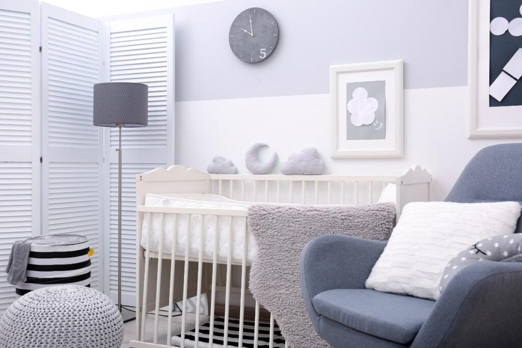 A cozy, modern nursery with a white crib, grey cushions shaped like clouds, a grey armchair with a white pillow, a fluffy grey blanket, a standing lamp, wall art, and a round wall clock. Soft, neutral tones decorate the room.