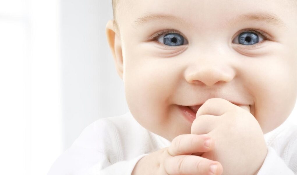 Close-up of a smiling baby with blue eyes and light hair, wearing a white outfit. The baby is looking at the camera with hands near their mouth, appearing happy and playful. The background is softly blurred and bright.
