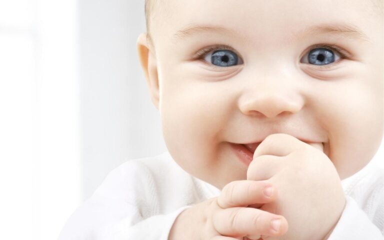 Close-up of a smiling baby with blue eyes and light hair, wearing a white outfit. The baby is looking at the camera with hands near their mouth, appearing happy and playful. The background is softly blurred and bright.