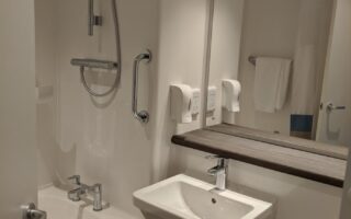 A compact, modern hotel bathroom with white walls. It features a bathtub with a mounted shower and grab bar, a small sink under a large mirror, a towel rack, two soap dispensers, and a neatly folded white towel on the tub.