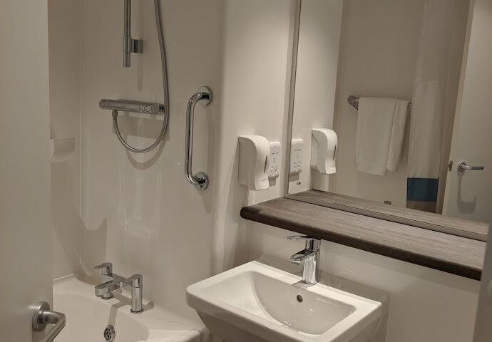A compact, modern hotel bathroom with white walls. It features a bathtub with a mounted shower and grab bar, a small sink under a large mirror, a towel rack, two soap dispensers, and a neatly folded white towel on the tub.