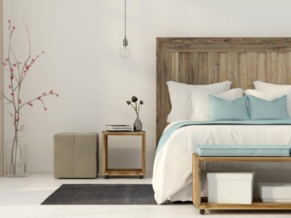 A modern bedroom with a rustic wooden headboard, white bedding, and light blue pillows. A wooden nightstand holds books, a vase, and a hanging bulb. A grey rug, beige ottoman, and minimalist decor complete the cozy, serene space.