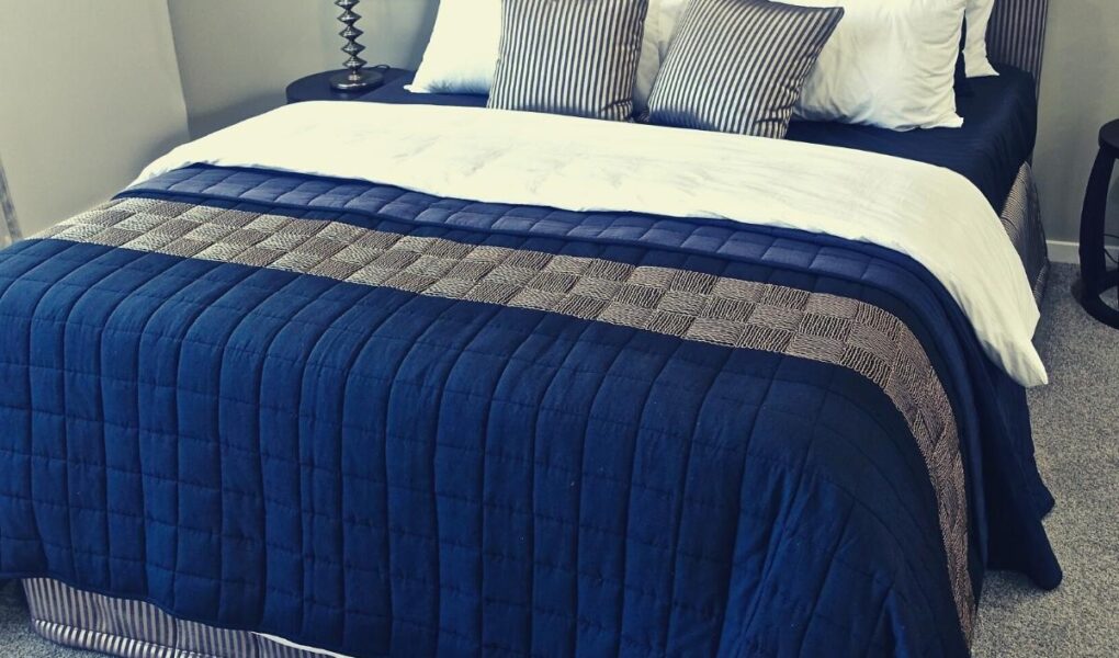 A neatly made bed with a blue quilt, white sheets, and multiple pillows in white, blue, and striped patterns. The headboard has vertical stripes, and there is a black lamp on a bedside table against a light-colored wall and carpeted floor.