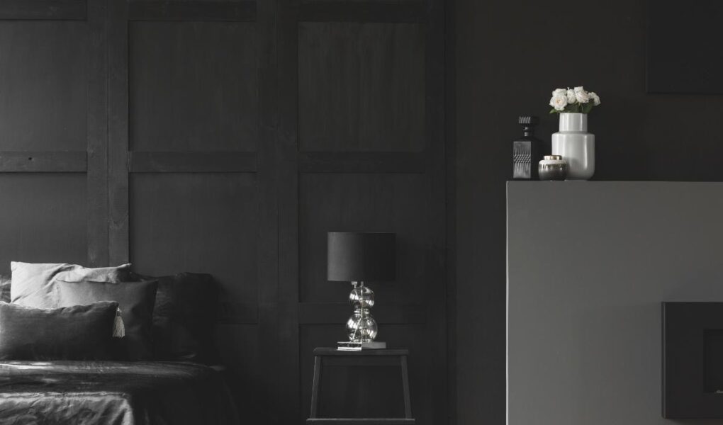 A modern, minimalist bedroom with dark walls, a bed with black linens and pillows, a black nightstand with a silver lamp, and a gray mantel holding white vases with flowers and candles. The space has a sleek, monochromatic look.