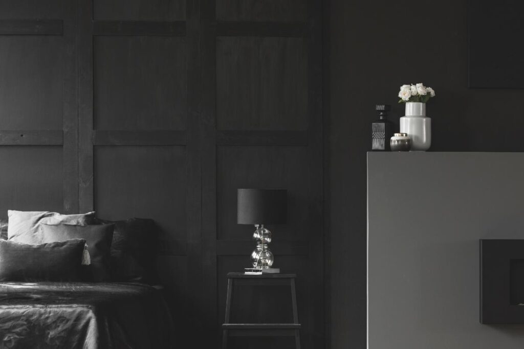 A modern, minimalist bedroom with dark walls, a bed with black linens and pillows, a black nightstand with a silver lamp, and a gray mantel holding white vases with flowers and candles. The space has a sleek, monochromatic look.