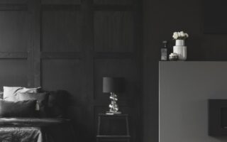 A modern, minimalist bedroom with dark walls, a bed with black linens and pillows, a black nightstand with a silver lamp, and a gray mantel holding white vases with flowers and candles. The space has a sleek, monochromatic look.