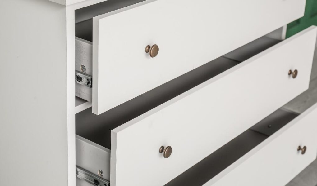 A close-up of a white wooden dresser with three drawers, two of which are partially open. The drawers have round bronze knobs, and the metal drawer slides are visible on the insides. The background is out of focus.