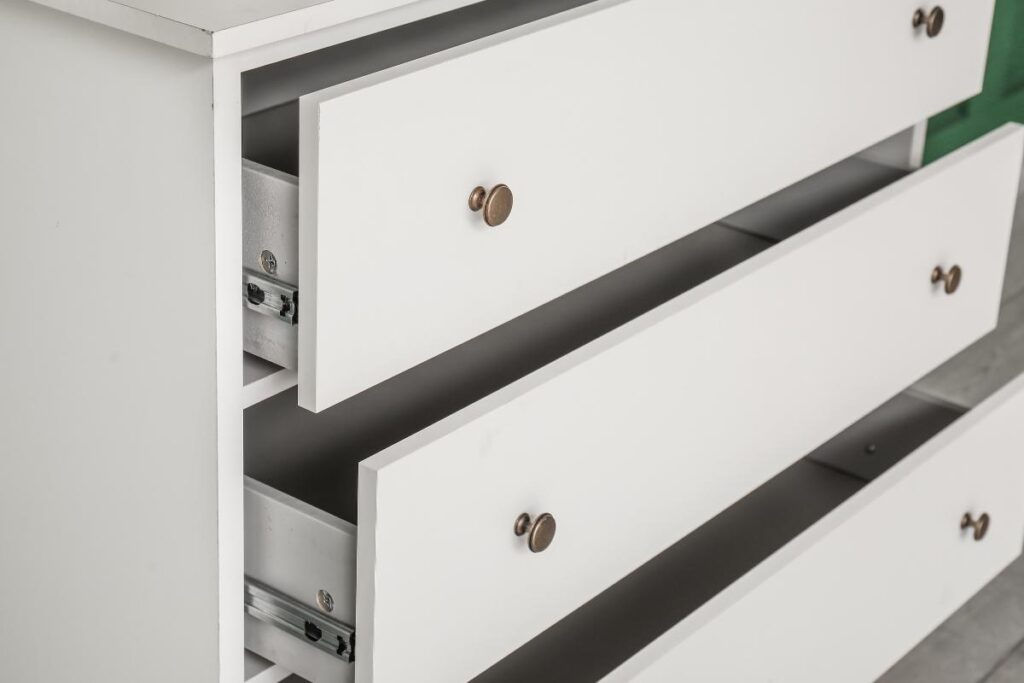A close-up of a white wooden dresser with three drawers, two of which are partially open. The drawers have round bronze knobs, and the metal drawer slides are visible on the insides. The background is out of focus.