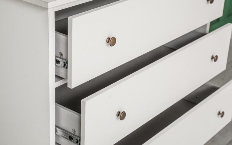 A close-up of a white wooden dresser with three drawers, two of which are partially open. The drawers have round bronze knobs, and the metal drawer slides are visible on the insides. The background is out of focus.