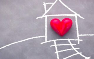 A simple chalk drawing of a house and path is on a gray surface. At the center of the house is a three-dimensional red heart, symbolizing love at home. A chalk-drawn smoke cloud rises from the house roof’s chimney.