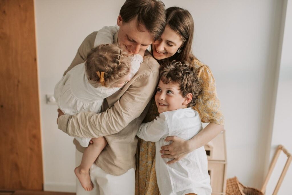 A family of four hugs closely indoors. A woman and a man smile, each holding a young child. The girl, with a bow in her hair, is held by the man, while a curly-haired boy hugs the man’s waist. All share a joyful, affectionate moment.