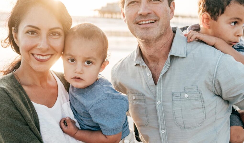 A smiling family of four stands on a beach at sunset. The woman holds a young child, the man stands beside her holding another child on his shoulder. They all wear casual clothes. The sun shines behind them, creating a warm, glowing light.