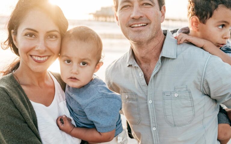 A smiling family of four stands on a beach at sunset. The woman holds a young child, the man stands beside her holding another child on his shoulder. They all wear casual clothes. The sun shines behind them, creating a warm, glowing light.