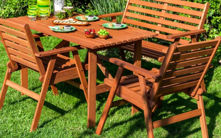A wooden outdoor dining set with a table, two chairs, and a bench sits on green grass. The table holds plates of food, a vase of white flowers, a pitcher, glasses, and snacks. Lush green bushes and plants are in the background.