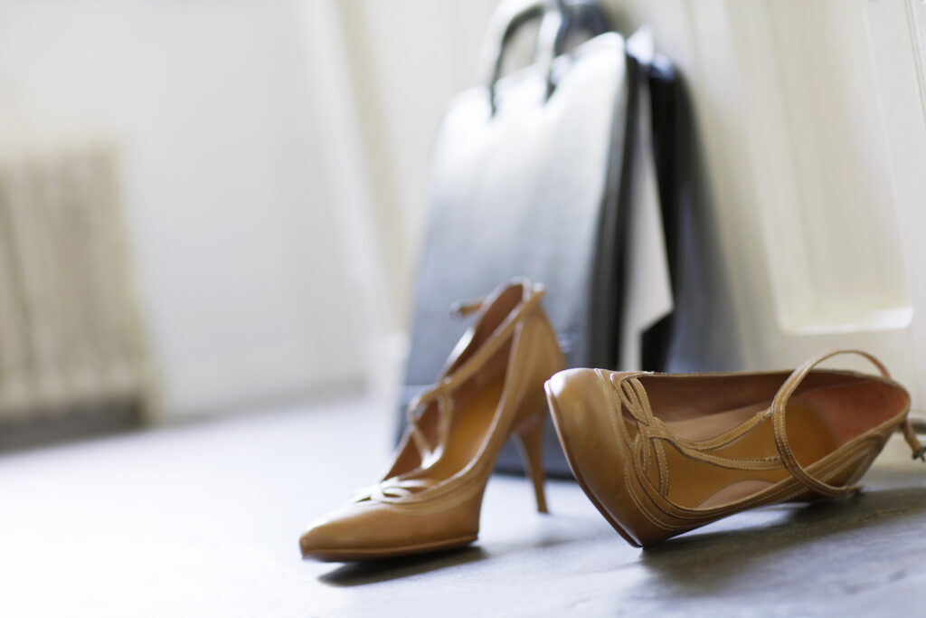 A pair of tan high-heeled shoes are placed on a gray floor near a white wall. In the blurred background, a black leather briefcase leans against the wall, suggesting a professional or office setting.