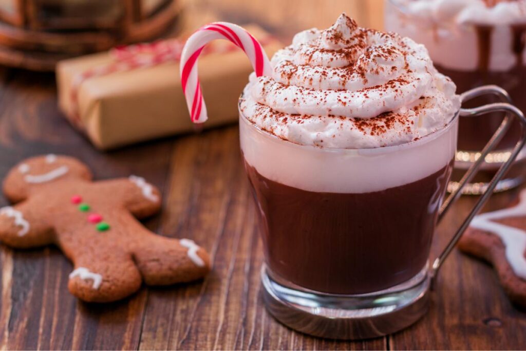 A glass mug of hot chocolate topped with whipped cream, cocoa powder, and a small candy cane sits on a wooden table. Next to it are a gingerbread cookie shaped like a person and a wrapped gift, evoking a festive holiday atmosphere.