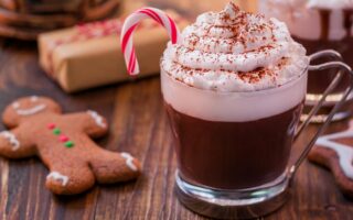 A glass mug of hot chocolate topped with whipped cream, cocoa powder, and a small candy cane sits on a wooden table. Next to it are a gingerbread cookie shaped like a person and a wrapped gift, evoking a festive holiday atmosphere.