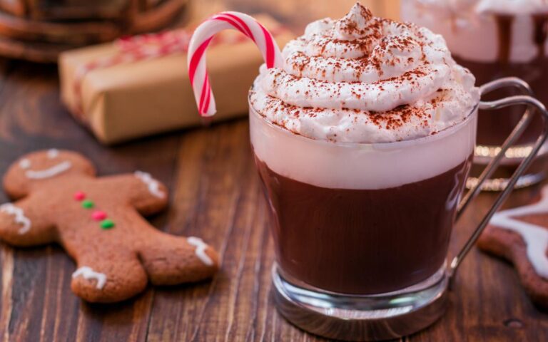 A glass mug of hot chocolate topped with whipped cream, cocoa powder, and a small candy cane sits on a wooden table. Next to it are a gingerbread cookie shaped like a person and a wrapped gift, evoking a festive holiday atmosphere.