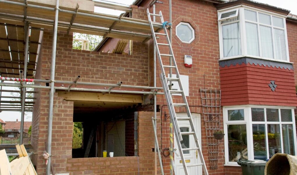 A brick house is under construction with scaffolding, ladders, and building materials outside. An extension is being added to the side, with an exposed wall and open window space. Tools, wood, and a concrete mixer are scattered in the front yard.