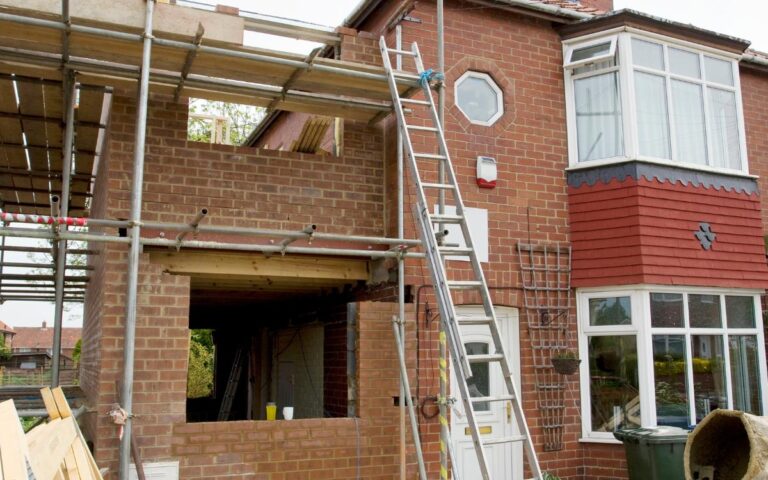 A brick house is under construction with scaffolding, ladders, and building materials outside. An extension is being added to the side, with an exposed wall and open window space. Tools, wood, and a concrete mixer are scattered in the front yard.