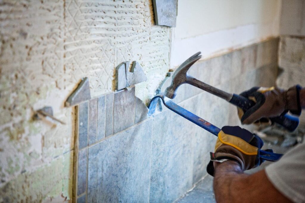 A person wearing gloves uses a hammer and a crowbar to remove ceramic wall tiles. The wall behind the tiles shows adhesive residue and partial damage. Some tiles are already removed, revealing the rough, textured surface underneath.