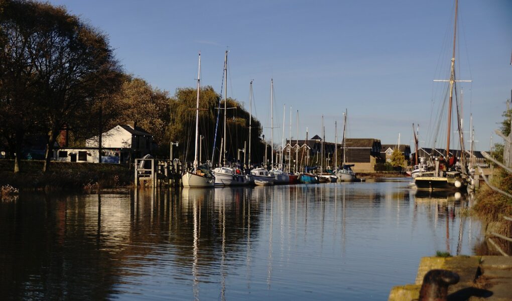 A peaceful marina with several sailboats docked along a calm river. Trees line the left bank and small buildings are visible in the background. The sky is clear and blue, and sunlight gently reflects off the water, creating a tranquil atmosphere.