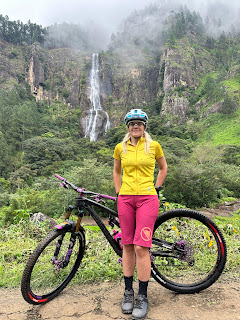 A woman in a yellow shirt, pink shorts, and a blue helmet stands smiling beside a black mountain bike. Behind her is lush greenery and a tall waterfall cascading down a forested cliff under a cloudy sky.