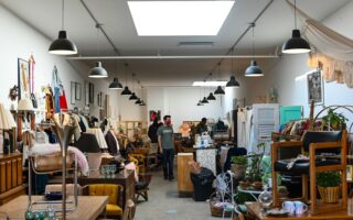 A brightly lit vintage shop with high ceilings displays eclectic furniture, lamps, clothes, and decor. Two people browse among the packed aisles. Art, hanging lights, and plants add charm to the cozy, organized space.