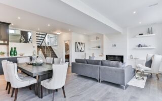 Modern open-concept living and dining area with light gray floors, a gray sectional sofa, an elegant dining table with eight white chairs, built-in shelves, decorative vases, wall art, and a staircase with glass railing in the background.