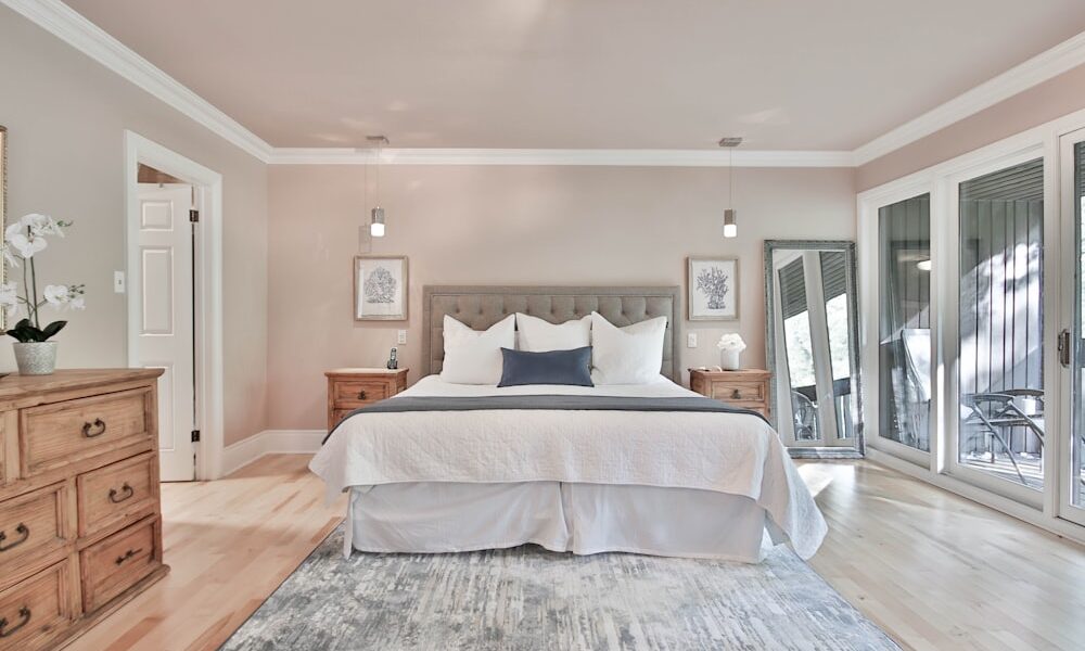 A bright bedroom with a large bed, white bedding, and gray headboard. Two wooden nightstands with lamps flank the bed. A mirror leans on the right wall near glass doors leading outside. Light wood floors and a patterned rug complete the neutral, airy decor.