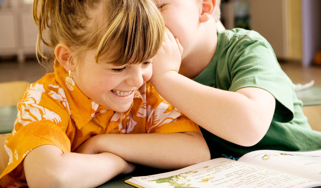 Two young children sit at a table with an open picture book. One child, smiling and wearing an orange shirt, listens as the other child, in a green shirt, leans in to whisper in their ear. The setting appears to be a classroom or library.