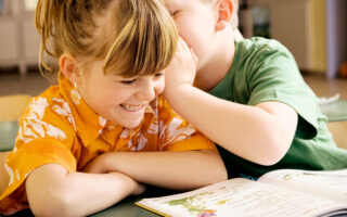 Two young children sit at a table with an open picture book. One child, smiling and wearing an orange shirt, listens as the other child, in a green shirt, leans in to whisper in their ear. The setting appears to be a classroom or library.