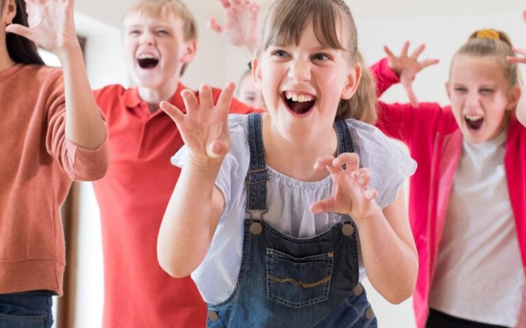 A group of four children enthusiastically pretend to be monsters, making claw gestures and playful faces. They are indoors, smiling and laughing, dressed in casual clothes. The background is bright with natural light coming through windows.