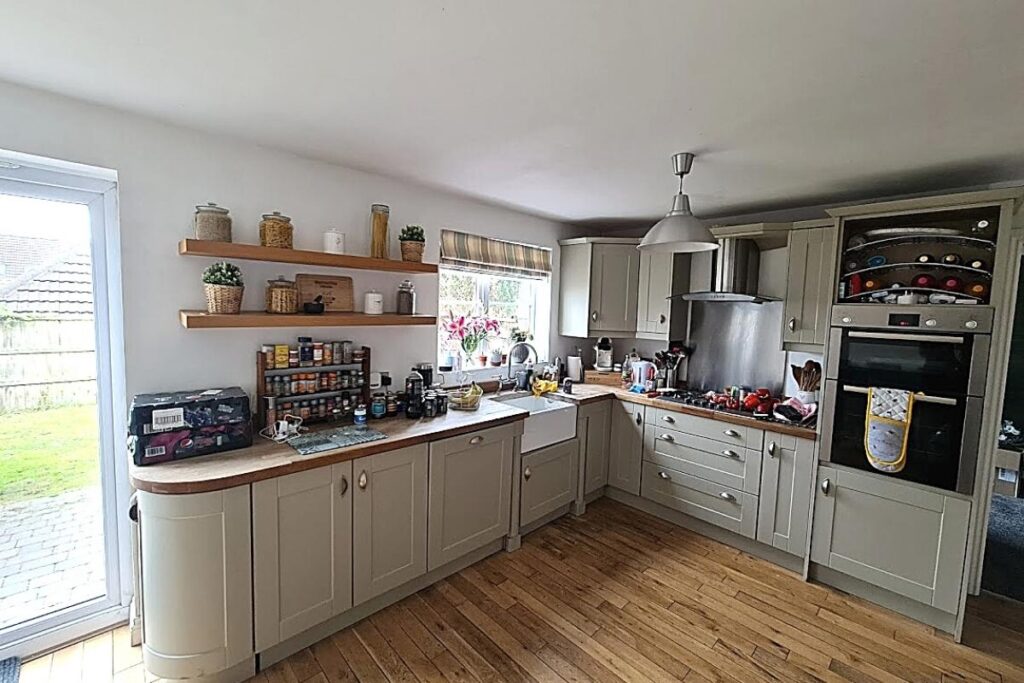 A bright, modern kitchen with light gray cabinets, wooden countertops, and wood flooring. Open shelves display jars and spices. The kitchen has a stove, oven, sink beneath a window, flowers, and various items on the counters. A glass door opens to a garden.