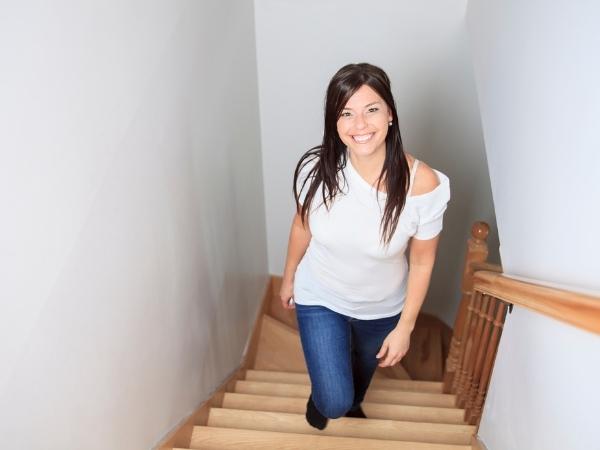 A woman with long brown hair, wearing a white top and blue jeans, is smiling and walking up a wooden staircase. The staircase has a wooden handrail, and the walls on either side are white, creating a bright, clean atmosphere.