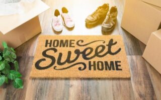 A doormat with the words "Home Sweet Home" sits on a wooden floor, surrounded by moving boxes, a pair of adult sneakers, children’s pink shoes, and a potted plant, all lit by warm sunlight streaming in.