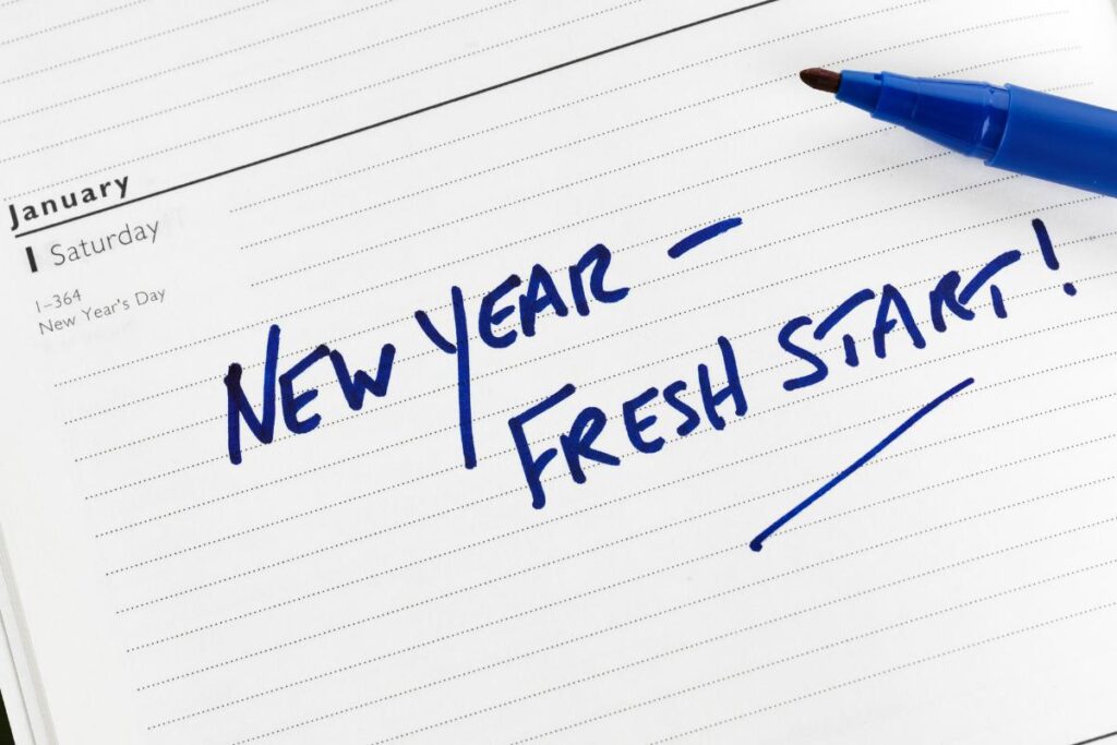 A blue marker rests on an open planner, showing a January Saturday. Written across the page in bold blue handwriting are the words “NEW YEAR – FRESH START!” The planner also notes “New Year’s Day” in the calendar entry.
