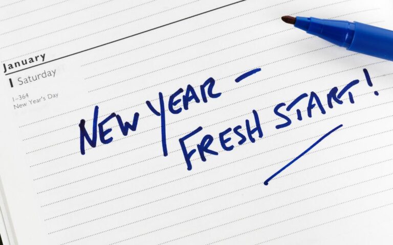 A blue marker rests on an open planner, showing a January Saturday. Written across the page in bold blue handwriting are the words “NEW YEAR – FRESH START!” The planner also notes “New Year’s Day” in the calendar entry.