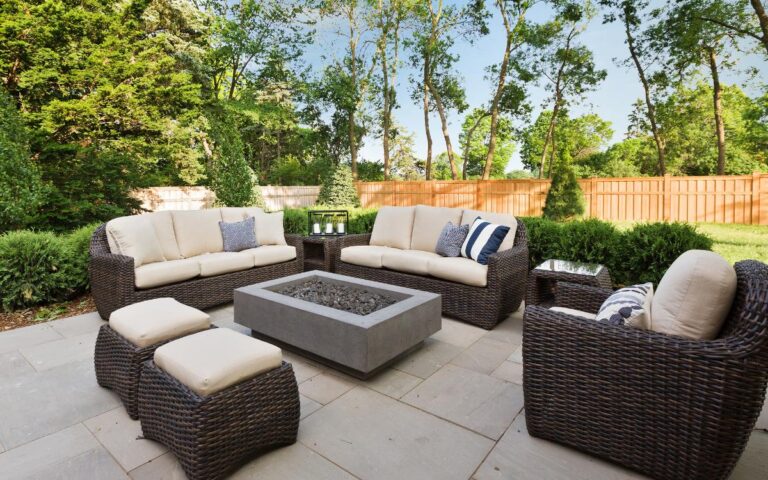 A patio seating area with wicker furniture topped with beige cushions surrounds a square fire pit. Two sofas, two armchairs, and two ottomans are arranged on gray stone tiles. The space is bordered by lush greenery and a wooden fence with tall trees behind it.