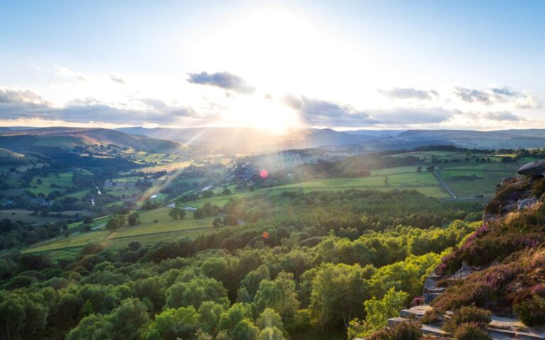 A scenic landscape shows rolling green hills, dense forests, and fields under a partly cloudy sky at sunset; sunlight beams spread across the view, illuminating the valley and casting soft shadows across the lush terrain.