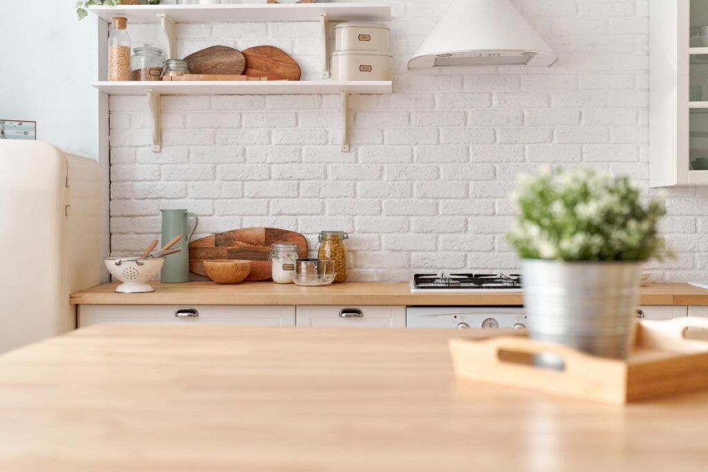 Bright, cozy small kitchen with white brick walls, wooden countertops, and shelves holding jars, bowls, and cutting boards. A plant in a metal pot sits on a wooden tray in the foreground, with a stove and white hood in the background.