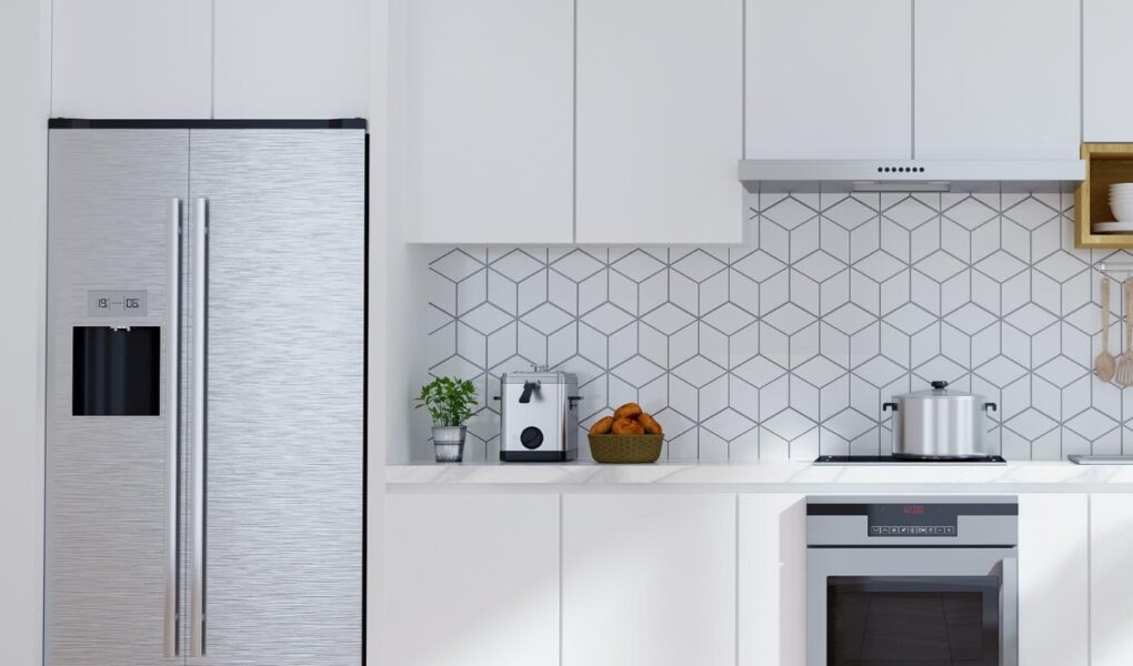 Modern small kitchen with white cabinets, stainless steel fridge, oven, and range hood. Counter holds a toaster, blender, and bowl of oranges. Geometric white tile backsplash and light wood herringbone floor keep the space clean and bright.