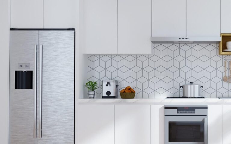 Modern small kitchen with white cabinets, stainless steel fridge, oven, and range hood. Counter holds a toaster, blender, and bowl of oranges. Geometric white tile backsplash and light wood herringbone floor keep the space clean and bright.