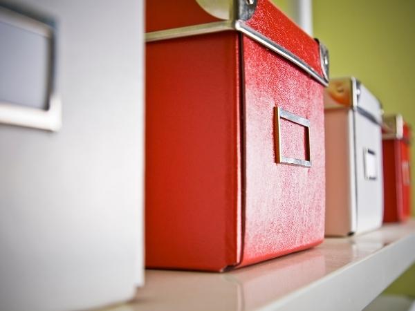 Close-up of colorful storage boxes with metal label holders, placed on a white shelf. The main focus is on a red box in the center, with parts of a white box on the left and a pink box on the right. Background is blurred and light green.