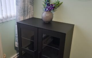 A small black cabinet with glass doors stands on a gray carpet in the corner of a room. A striped vase with purple and pink flowers sits on top. Nearby, sheer white curtains with a leafy pattern cover a window, and round shelves hang on the pale green wall.